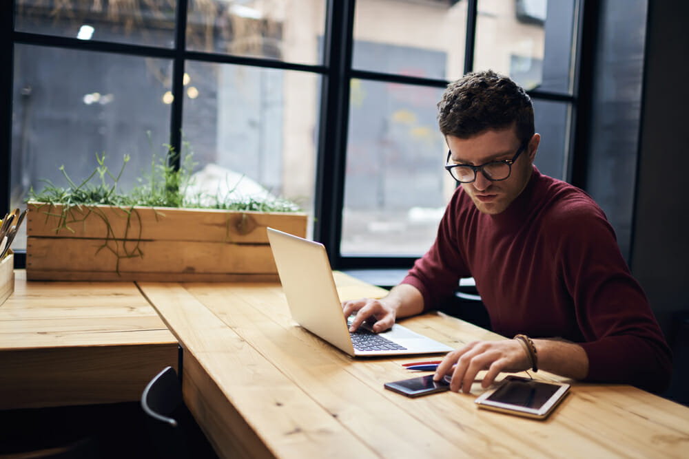 Man sitting at a wood table working on his laptop and phone