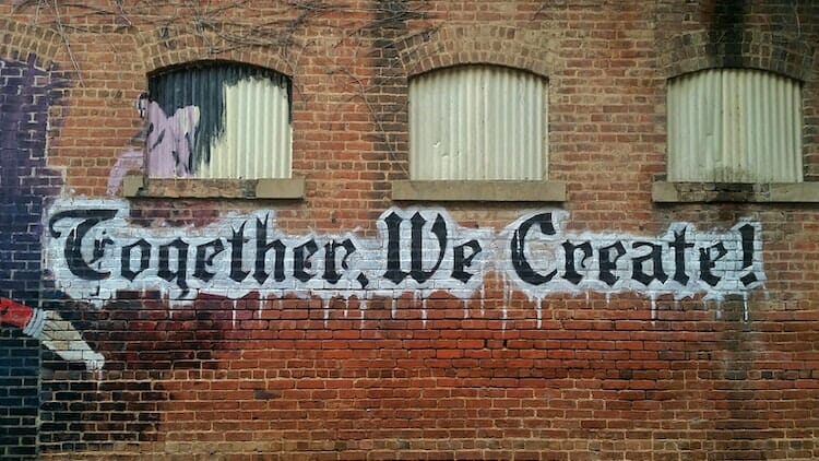 Photo of a brick wall with three covered windows. Graffiti says "Together, We Create!"