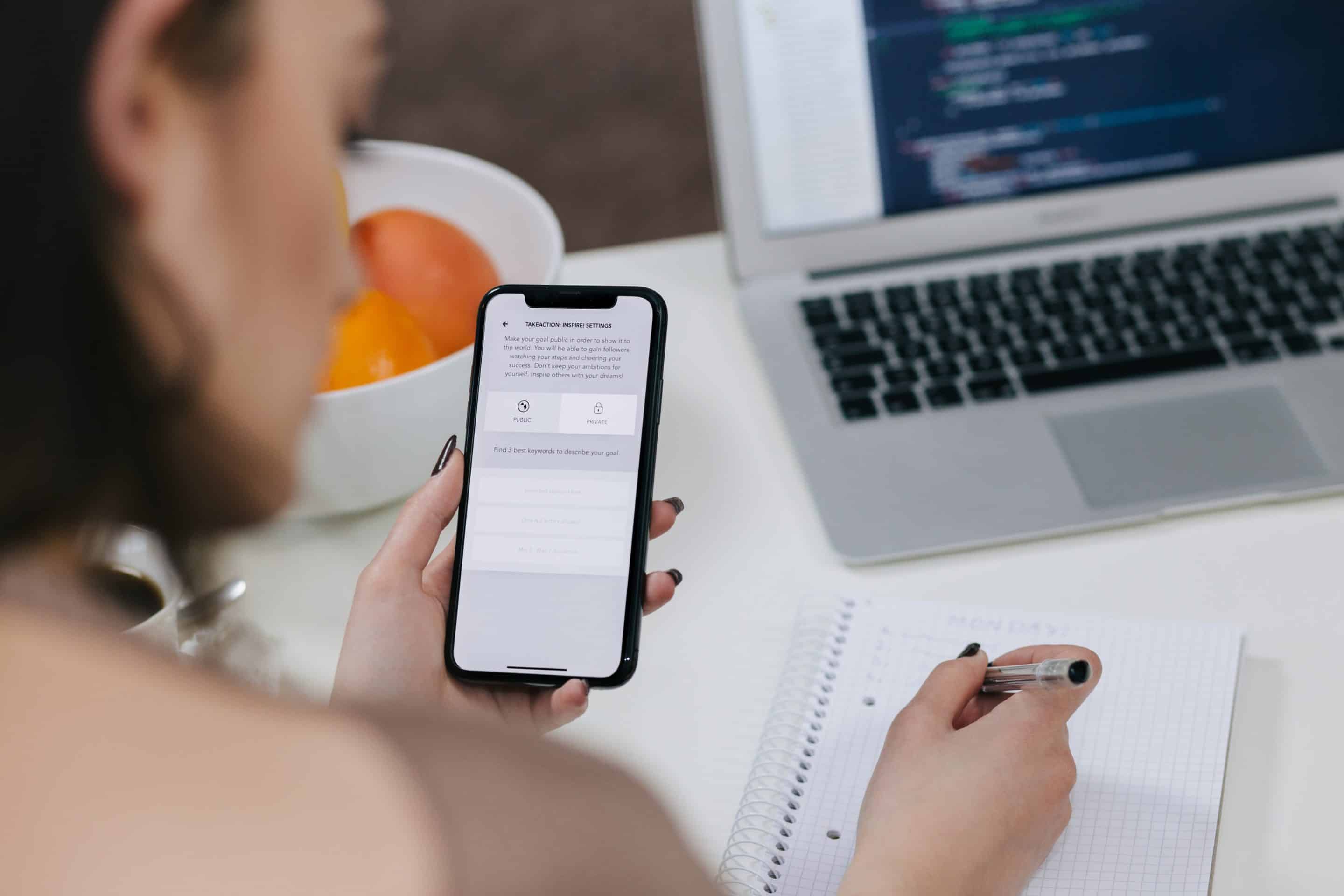 Woman holding her mobile phone in her left hand and writing in a notebook with her right hand.